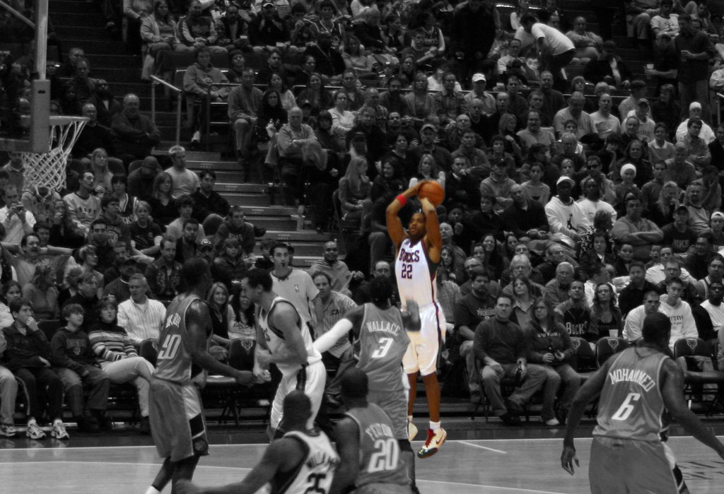 A player rises for a jump shot as defenders and a packed crowd look on